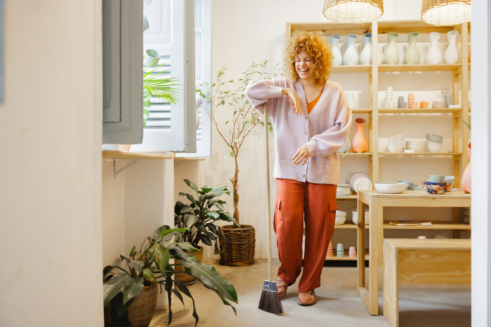 Woman sweeping a pottery shop beside shelves of ceramics